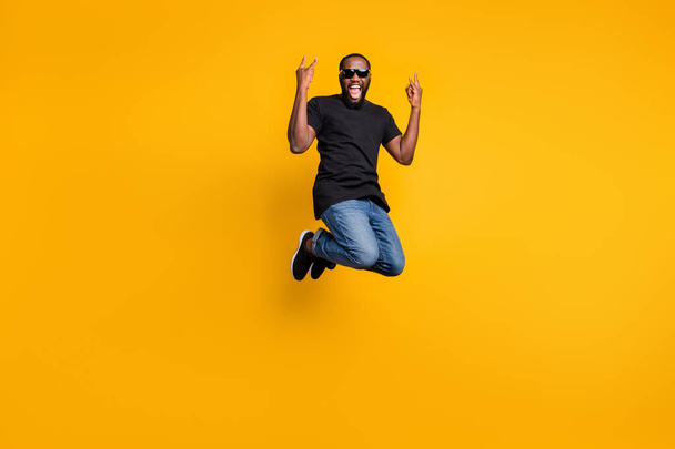 Full length photo of crazy funky afro american guy jump fun rock concert scream show horns wear youth style denim jeans isolated over yellow color backround - Foto, imagen