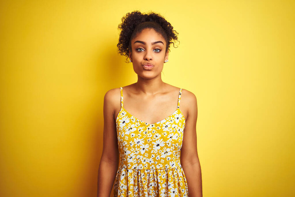 African american woman wearing casual floral dress standing over isolated yellow background looking at the camera blowing a kiss on air being lovely and sexy. Love expression. - Foto, imagen