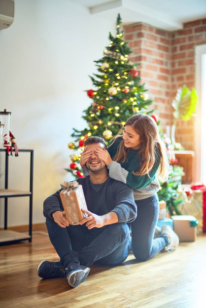 Jong mooi koppel glimlachend gelukkig en zelfverzekerd. vrouw bedekken ogen van man surpise haar met geschenk rond kerstboom thuis - Foto, afbeelding