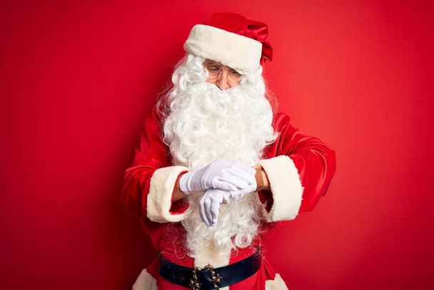 Orta yaşlı, yakışıklı, Noel Baba kostümü giymiş kırmızı arka planda duran rahat ve kendinden emin bir şekilde kol saatine bakıyor. - Fotoğraf, Görsel