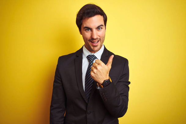 Νεαρός όμορφος επιχειρηματίας με κοστούμι και γραβάτα στέκεται πάνω από απομονωμένο κίτρινο φόντο κάνοντας χαρούμενη χειρονομία με το χέρι. Έγκριση έκφρασης κοιτάζοντας την κάμερα που δείχνει επιτυχία. - Φωτογραφία, εικόνα