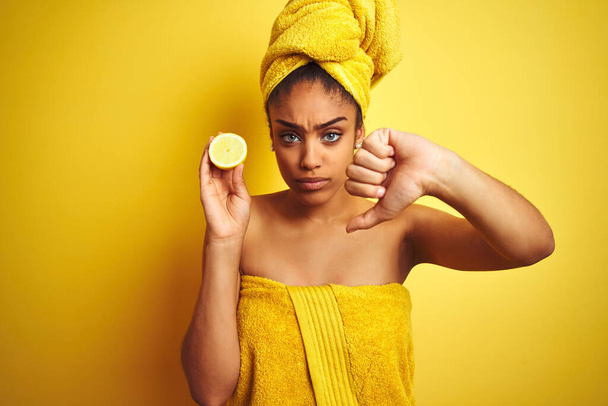 Mujer afro usando toalla después de la ducha sosteniendo rodaja de limón sobre fondo amarillo aislado con la cara enojada, signo negativo que muestra aversión con los pulgares hacia abajo, concepto de rechazo
 - Foto, imagen