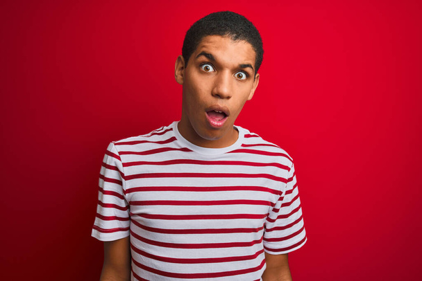 Mladý pohledný arabský muž v pruhovaném tričku přes izolované červené pozadí v šokující tváři, skeptický a sarkastický, překvapený s otevřenými ústy - Fotografie, Obrázek
