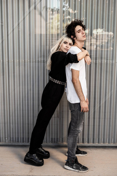 Vista lateral do casal na moda apoiando uns aos outros, enquanto a mulher se inclina no namorado de volta contra a parede listrada do edifício moderno
 - Foto, Imagem