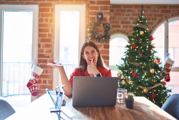 Beautiful woman sitting at the table working with laptop at home around christmas tree Showing palm hand and doing ok gesture with thumbs up, smiling happy and cheerful - Photo, Image