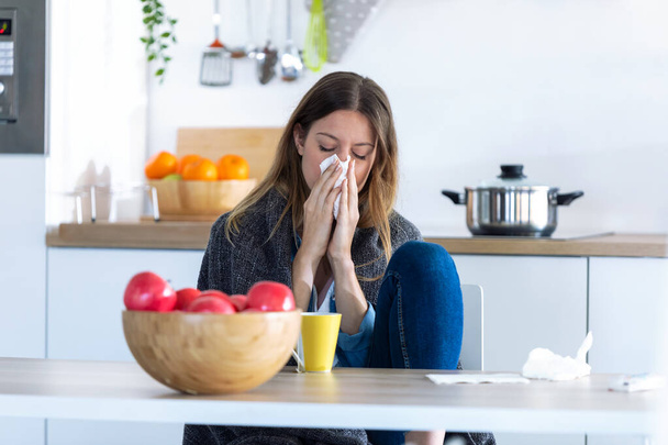 Illness young woman sneezing in a tissue while sitting in the kitchen at home. - Valokuva, kuva
