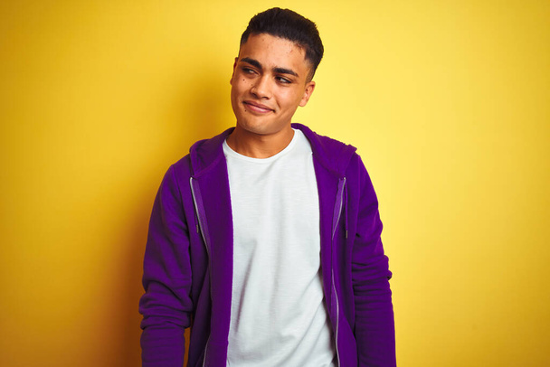 Молодий бразільський чоловік, одягнений у пурпурову сорочку, стоїть над жовтим фоном, усміхаючись, дивлячись убік і дивлячись убік,. - Фото, зображення