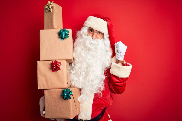 Старший чоловік у костюмі Санта Клауса тримає вежу подарунків над ізольованим червоним фоном, верещачи гордо і відзначаючи перемогу і успіх дуже збуджені, підбадьорюючи емоції. - Фото, зображення