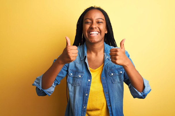 Mladá Afroameričanka v džínové košili, stojící nad osamělým žlutým nápisem úspěchu na pozadí, dělá pozitivní gesto rukou, palce nahoru, usmívá se a je šťastná. Veselý výraz a gesto vítěze. - Fotografie, Obrázek