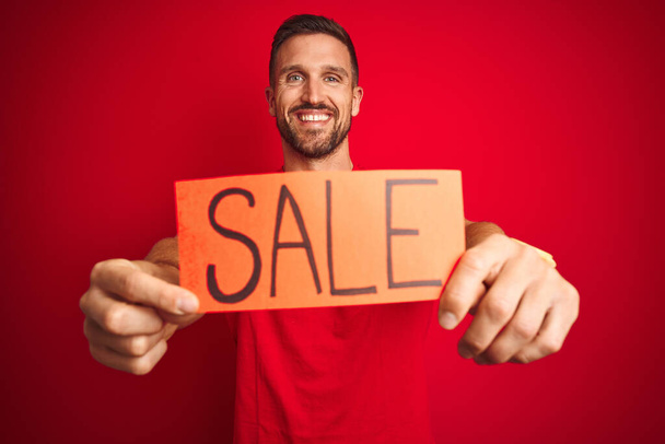 Young man holding sale advertising poster board over red isolated background with a happy face standing and smiling with a confident smile showing teeth - Fotoğraf, Görsel
