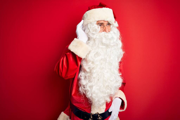 Keski-ikä komea mies yllään Santa puku seisoo yli eristetty punainen tausta hymyillen käsi korvan kuunnellen kuulo huhu tai juoruja. Kuurous-käsite
. - Valokuva, kuva