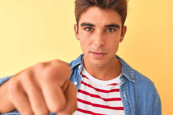 Νεαρός όμορφος άντρας με ριγέ t-shirt και denim πουκάμισο πάνω από το απομονωμένο κίτρινο φόντο που δείχνει με το δάχτυλο στην κάμερα και σε σας, σημάδι στο χέρι, θετική και σίγουρη χειρονομία από μπροστά - Φωτογραφία, εικόνα