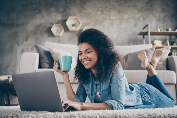 Photo of amazing dark skin curly lady browsing using notebook texting with friends drink hot beverage lying cozy floor near sofa wear casual denim outfit flat indoors - 写真・画像