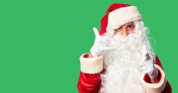 Middle age handsome man wearing Santa Claus costume and beard standing smiling doing talking on the telephone gesture and pointing to you. Call me. - 写真・画像