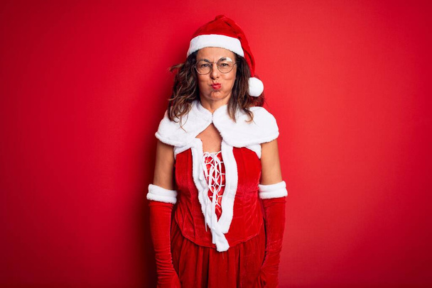 Piękna kobieta w średnim wieku ubrana w kostium Świętego Mikołaja na odizolowanym czerwonym tle nadyma policzki z zabawną twarzą. Usta napompowane powietrzem, szalony wyraz twarzy. - Zdjęcie, obraz