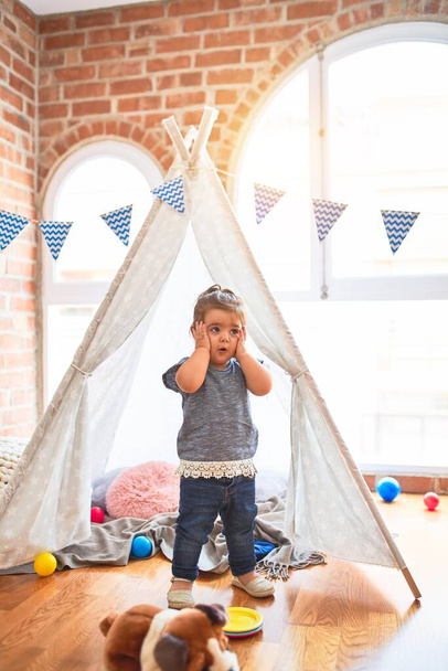 Beautiful toddler standing inside tipi with hands on face surprised and worried at kindergarten - Zdjęcie, obraz