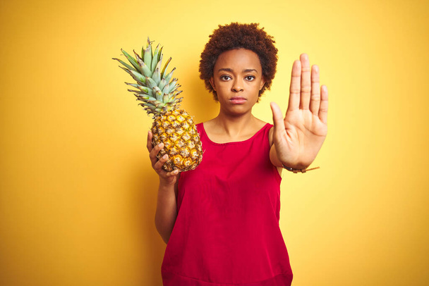 Afroamerykanka trzymając tropikalny ananas nad żółtym odizolowanym tle z otwartą ręką robi znak stop z poważnym i pewnym siebie wyrazem twarzy, gest obrony - Zdjęcie, obraz