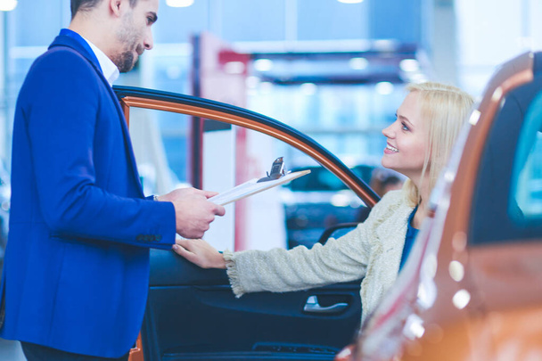 Beautiful young woman is talking to handsome car dealership worker while choosing a car in dealership - Fotó, kép