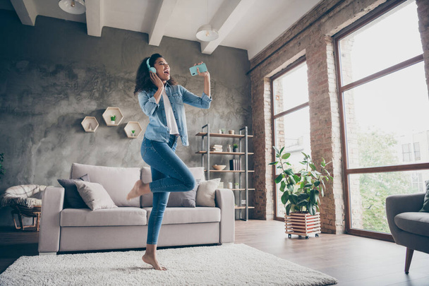 Full body photo of beautiful dark skin wavy lady holding telephone like microphone wear modern earflaps singing sitting floor near couch casual denim outfit living room indoors - Fotoğraf, Görsel