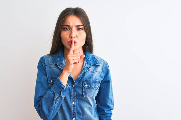 Young beautiful woman wearing casual denim shirt standing over isolated white background asking to be quiet with finger on lips. Silence and secret concept. - 写真・画像