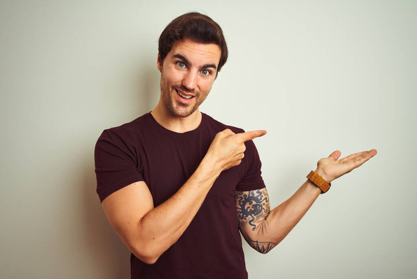 Молодий красивий чоловік з татуюванням носить фіолетову повсякденну футболку над ізольованим білим тлом, здивований і посміхається камері, представляючи рукою і вказуючи пальцем
. - Фото, зображення
