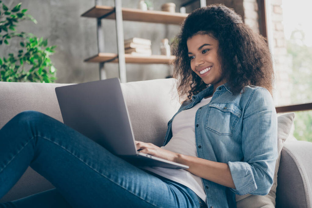 Closeup profile photo of pretty amazed dark skin curly lady notebook on knees texting friends read cool good news lying comfy couch casual denim outfit living room indoors - 写真・画像