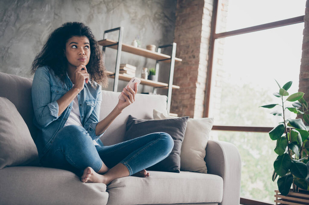 Photo of pretty dark skin curly lady hold telephone deeply minded think over new port text finger on chin sitting comfy couch casual denim outfit living room indoors - Foto, Imagem