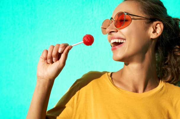 Close up portrait of a young stylish laughing emotional woman with big red lollipop standing against blue background. - Valokuva, kuva