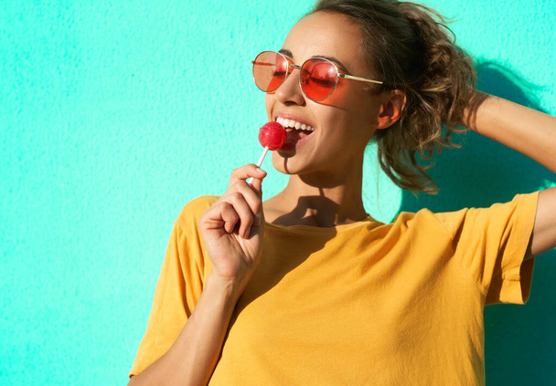 young stylish smiling woman standing against blue background and joyfully licking a big red lollipop. - Photo, image