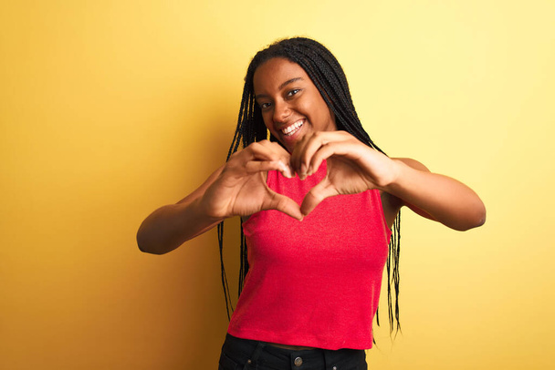 Femme afro-américaine portant un t-shirt décontracté rouge debout sur un fond jaune isolé souriant en amour faisant forme de symbole de coeur avec les mains. Concept romantique
. - Photo, image