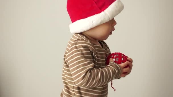 Lindo bebé en un sombrero de Santa lleva una bola roja de Navidad en sus manos. Preparándose para la noche de Navidad, feliz Noel
 - Imágenes, Vídeo