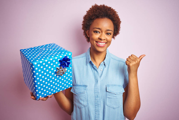 Afrika kökenli Amerikalı bir kadın pembe izole edilmiş arka planda doğum günü hediyesini tutuyor ve gülümseyen bir gülümsemeyle baş parmağıyla işaret ediyor. - Fotoğraf, Görsel