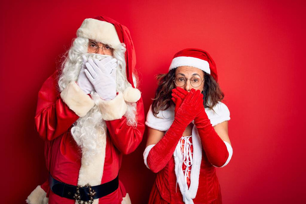 Couple d'âge moyen portant costume de Père Noël et des lunettes sur fond rouge isolé choqué couvrant la bouche avec les mains pour erreur. Concept secret
. - Photo, image