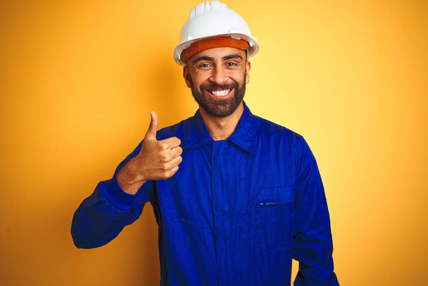 Komea intialainen työntekijä mies yllään yhtenäinen ja kypärä yli eristetty keltainen tausta tekee onnellinen peukut ylös ele käsin. Hyväksymällä ilme katsomalla kameraa osoittaa menestystä
. - Valokuva, kuva