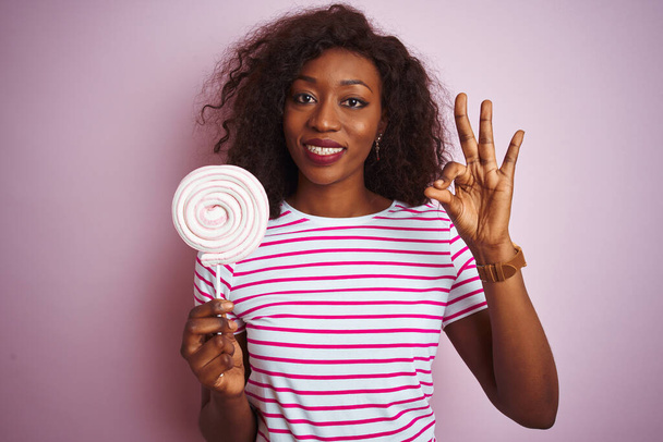 Nuori afrikkalainen amerikkalainen nainen syö makeaa karkkia seisoo eristetty vaaleanpunainen tausta tekee ok merkki sormilla, erinomainen symboli
 - Valokuva, kuva