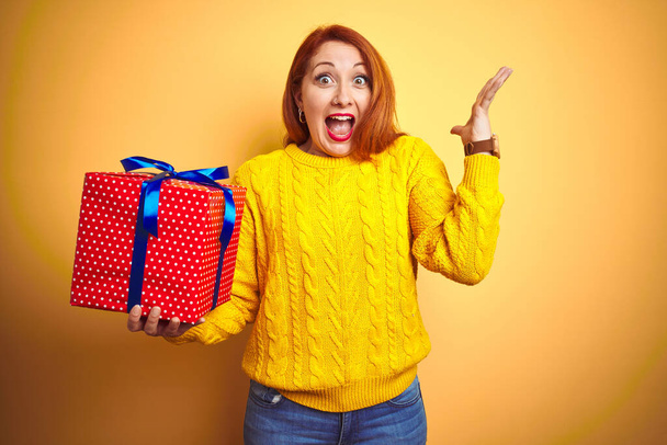 Young beautiful redhead woman holding birthday gift over yellow isolated background very happy and excited, winner expression celebrating victory screaming with big smile and raised hands - Фото, изображение