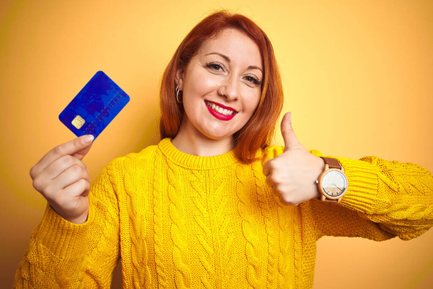 Jovem cliente ruiva mulher segurando cartão de crédito sobre amarelo isolado fundo feliz com grande sorriso fazendo ok sinal, polegar para cima com os dedos, excelente sinal
 - Foto, Imagem