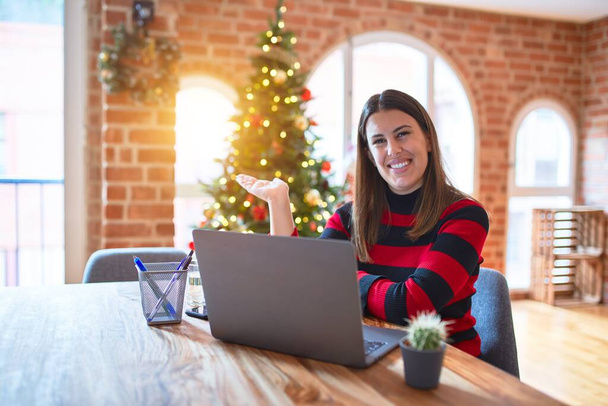 クリスマスツリーの周りの自宅でノートパソコンで作業テーブルに座っている美しい女性は陽気なプレゼンテーションを笑顔とカメラを見て手の手のひらで指して. - 写真・画像
