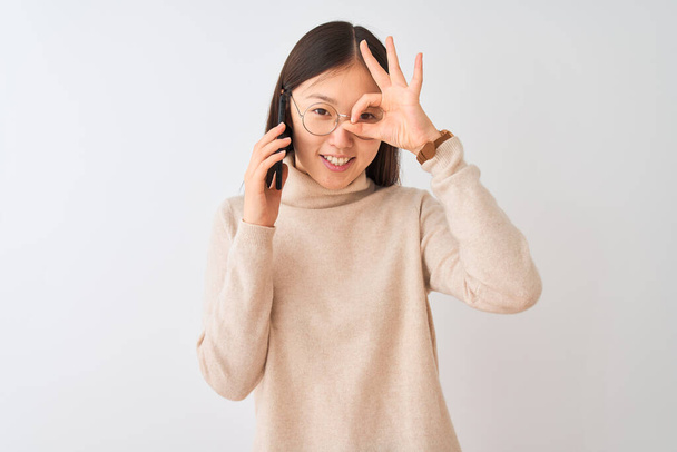 Молодая китаянка говорит по смартфону на изолированном белом фоне делает хорошо жест с улыбкой на руке, глаза смотрят сквозь пальцы с счастливым лицом
. - Фото, изображение