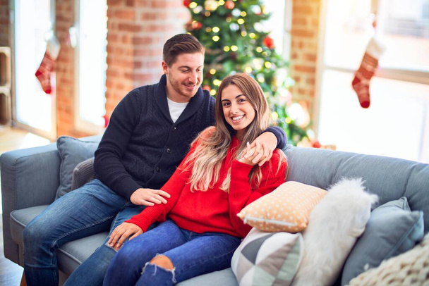 Young beautiful couple smiling happy and confident. Sitting on the sofa hugging around christmas tree at home - Φωτογραφία, εικόνα