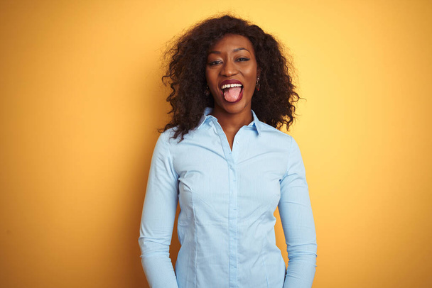 Femme d'affaires afro-américaine portant une chemise élégante sur fond jaune isolé langue collante heureux avec une expression drôle. Concept d'émotion
. - Photo, image