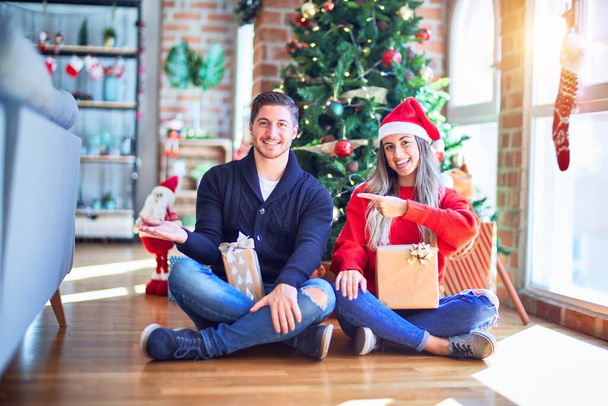 Mladý pár nosí Santa Claus klobouk sedí na podlaze kolem vánočního stromečku doma ohromený a usmívá se do kamery, zatímco prezentuje s rukou a ukazuje prstem. - Fotografie, Obrázek