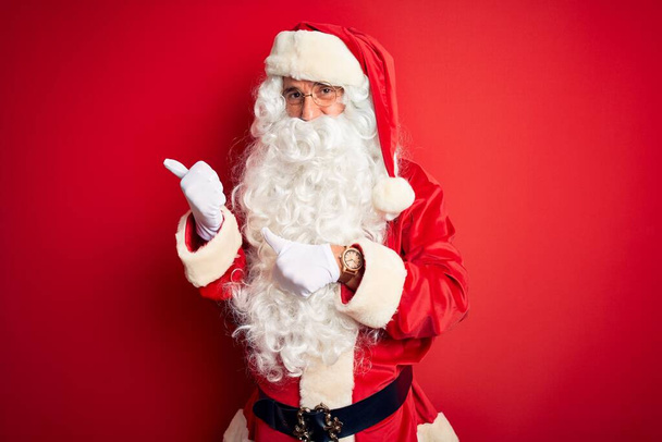 Keski-ikä komea mies yllään Santa puku seisoo yli eristetty punainen tausta osoittaa takana käsi ja peukalot pystyssä, hymyillen luottavainen
 - Valokuva, kuva