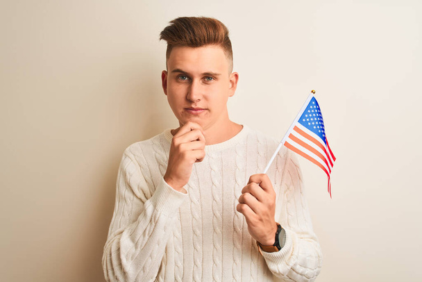 Joven hombre guapo sosteniendo bandera de EE.UU. sobre fondo blanco aislado cara seria pensando en la pregunta, idea muy confusa
 - Foto, Imagen