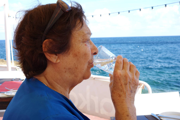 Porträt einer älteren Frau, die Wasser aus einem Glas trinkt. Schuss im Freien am Meer - Foto, Bild