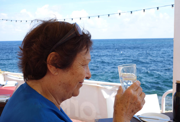 Profilo ritratto di una donna anziana che beve acqua da un bicchiere. Girato all'aperto in riva al mare
 - Foto, immagini