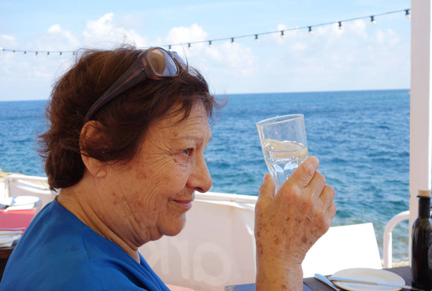 Perfil retrato de uma mulher idosa bebendo água de um copo. Tiro ao ar livre pelo mar
 - Foto, Imagem