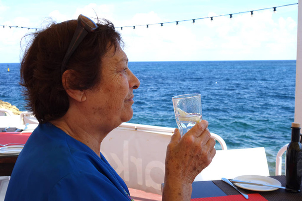 Porträt einer älteren Frau, die Wasser aus einem Glas trinkt. Schuss im Freien am Meer - Foto, Bild