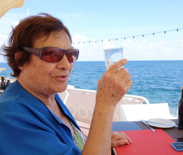 年配の女性がガラスから水を飲んでいる。海の外で撃たれた - 写真・画像