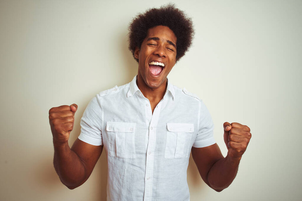 Африканський американець з волоссям афроамериканського кольору, що стоїть над білим фоном, дуже щасливий і захоплений жестом переможця з піднятими руками, посміхаючись і кричачи про успіх. Святкування.. - Фото, зображення